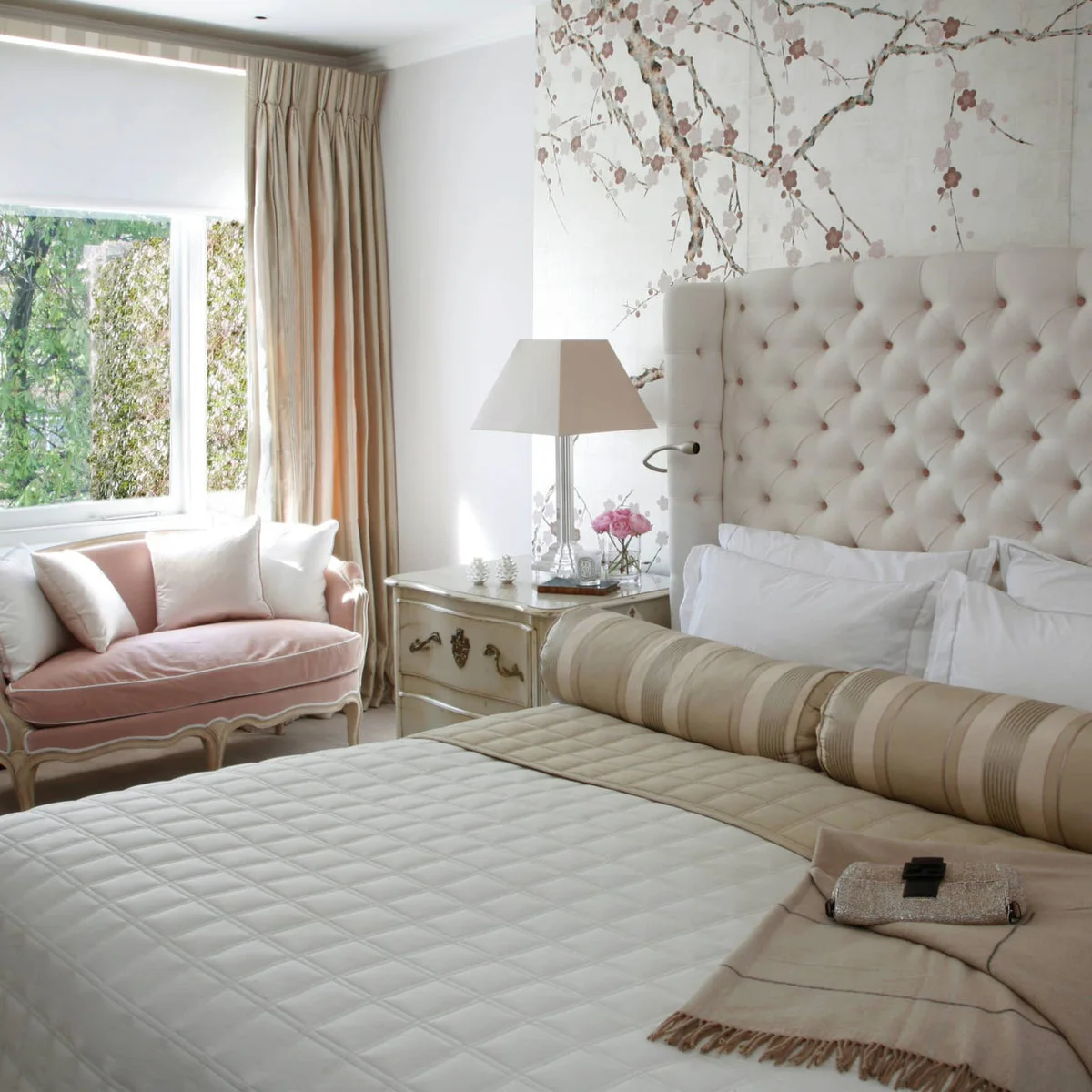 Pale tufted headboard with blossom-patterned wall covering and a blush settee near the window