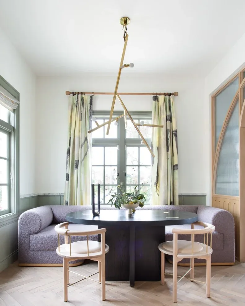 Purple upholstered banquette with curved arms behind a dark round table, with pale wooden chairs, patterned curtains, gray-green lower walls, and a branching light.