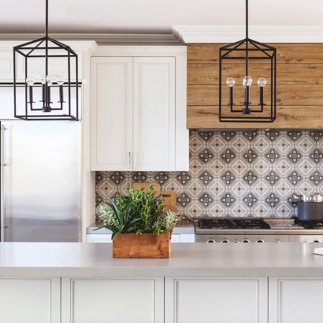 A horizontal timber hood sits above patterned backsplash tiles between white cabinets and black lantern pendants.