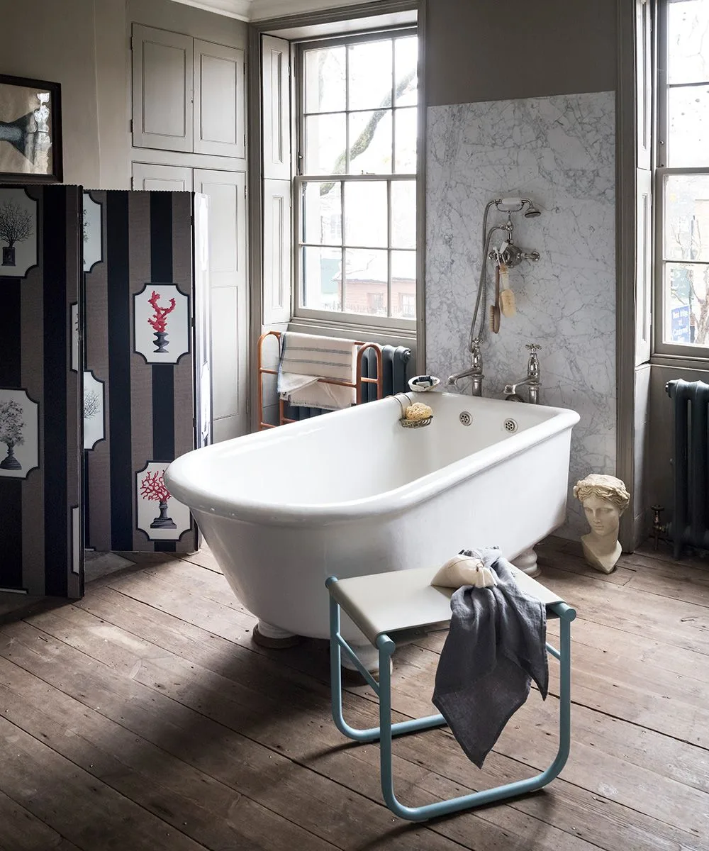 White freestanding tub beside tall sash windows, a dark decorative folding screen and gray upper walls.
