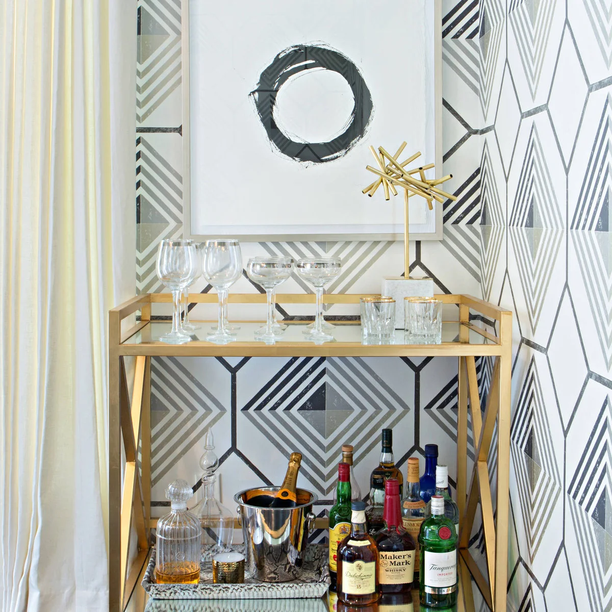 Gold-toned two-tier drinks stand with glassware above bottles beside geometric wallpaper and a curtain