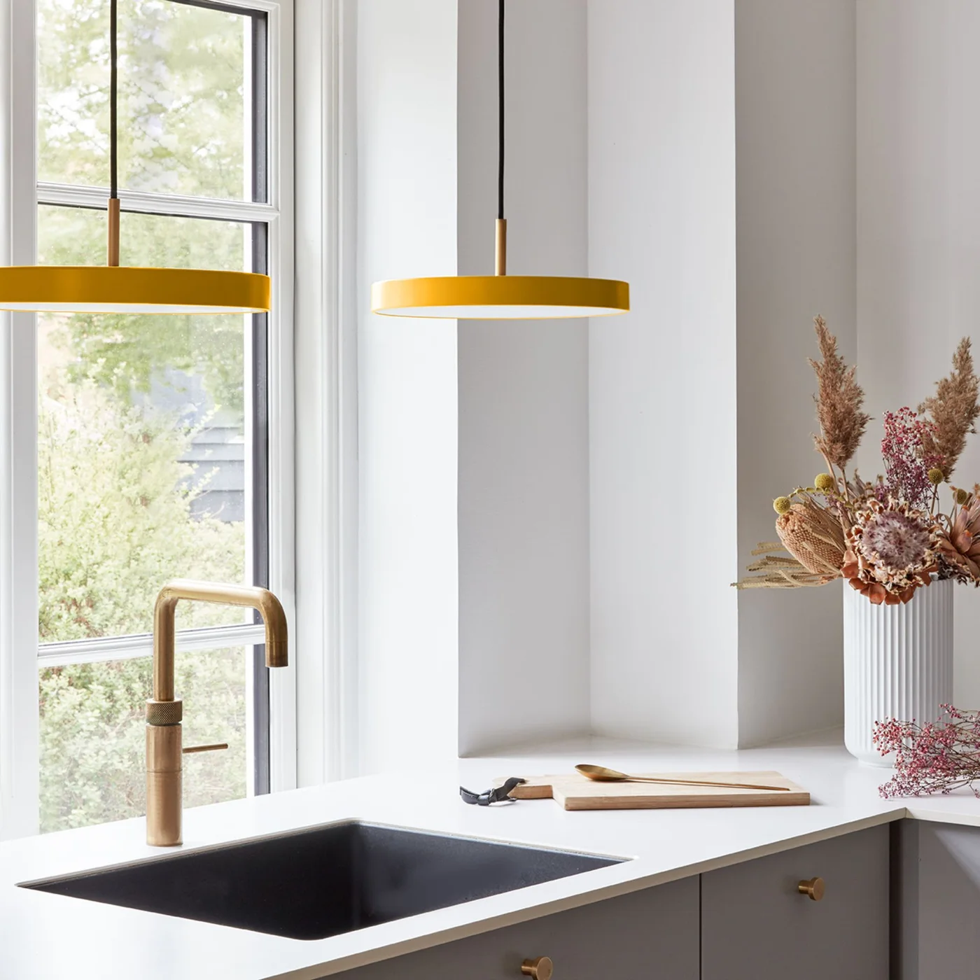 Two mustard-yellow pendants hang beside a window over a dark sink and gold-colored tap