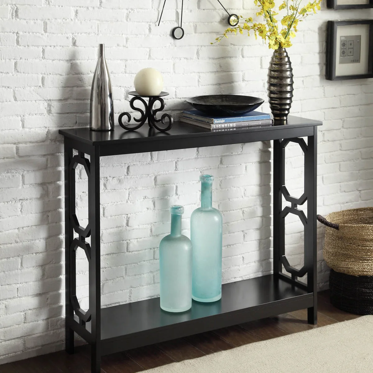 A black console against white brick with tall vessels, a shallow bowl on books and pale blue bottles below.