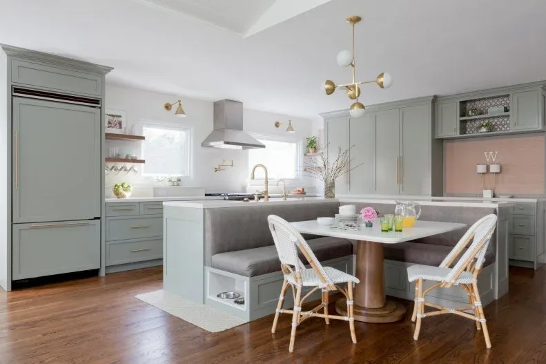 Gray upholstered banquette against a pale kitchen peninsula, with a white table on a gold-colored cylindrical base, woven chairs, gray cabinets, and an open lower cubby.