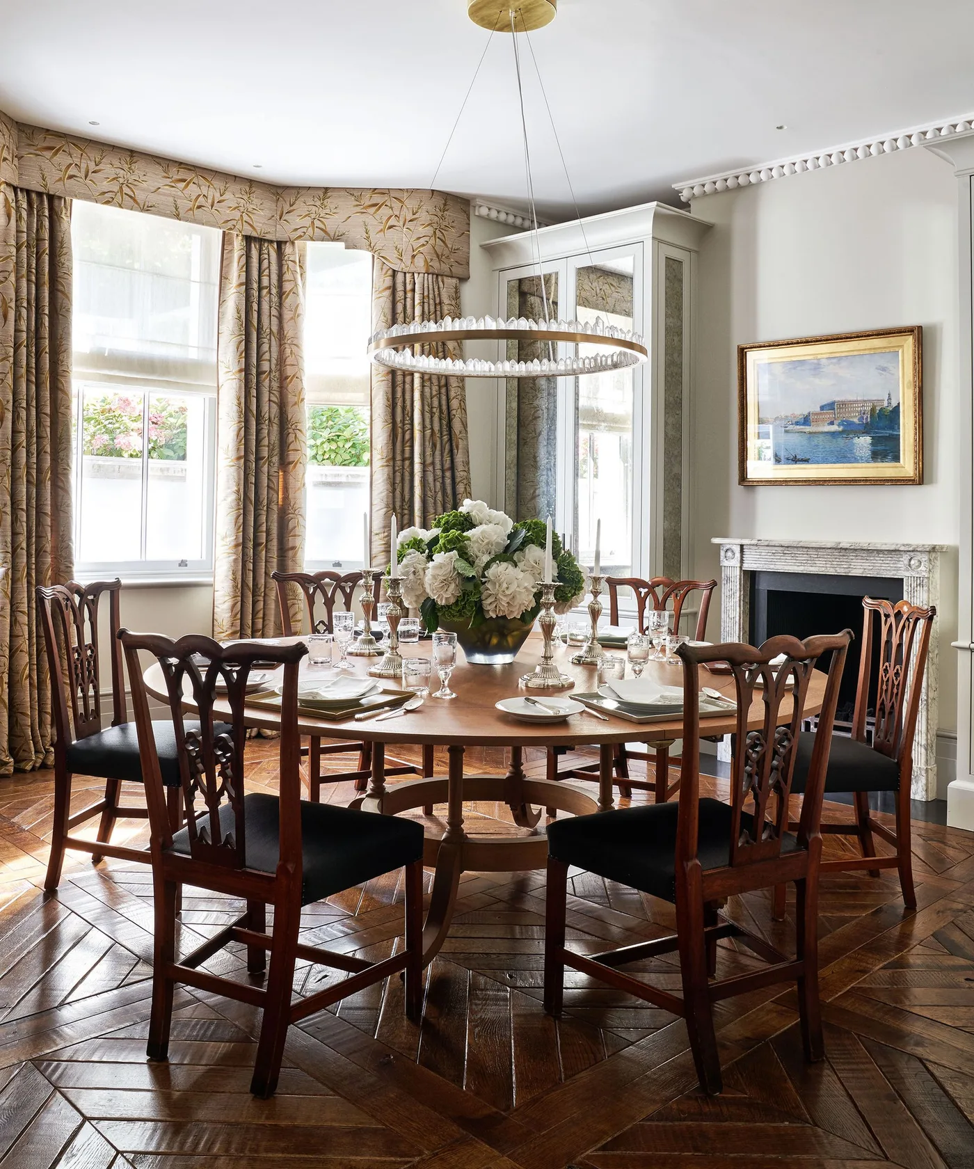 Country dining room with a large round wooden table, shaped wooden chairs with dark seats, patterned curtains and a circular hanging light.