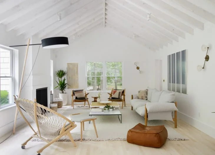 A white vaulted living room has a pale sofa, wood-framed chairs, a woven lounge chair, a glass coffee table and a brown floor cushion