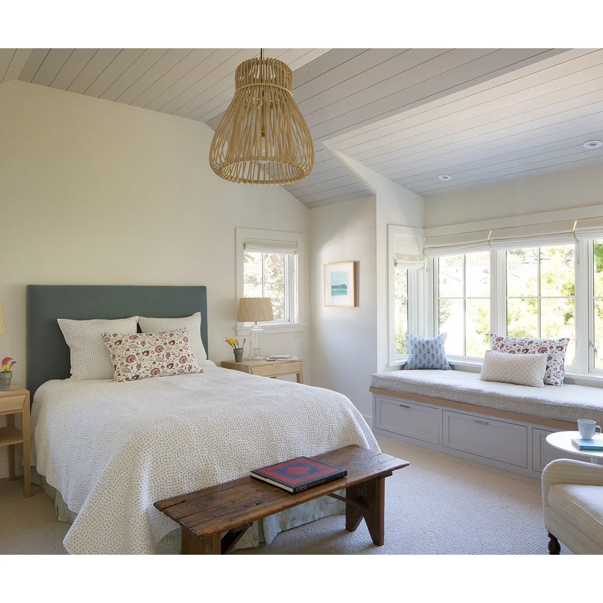 A gray cushioned window seat with drawers beside a bed with a blue headboard, pale bedding and a wooden bench.