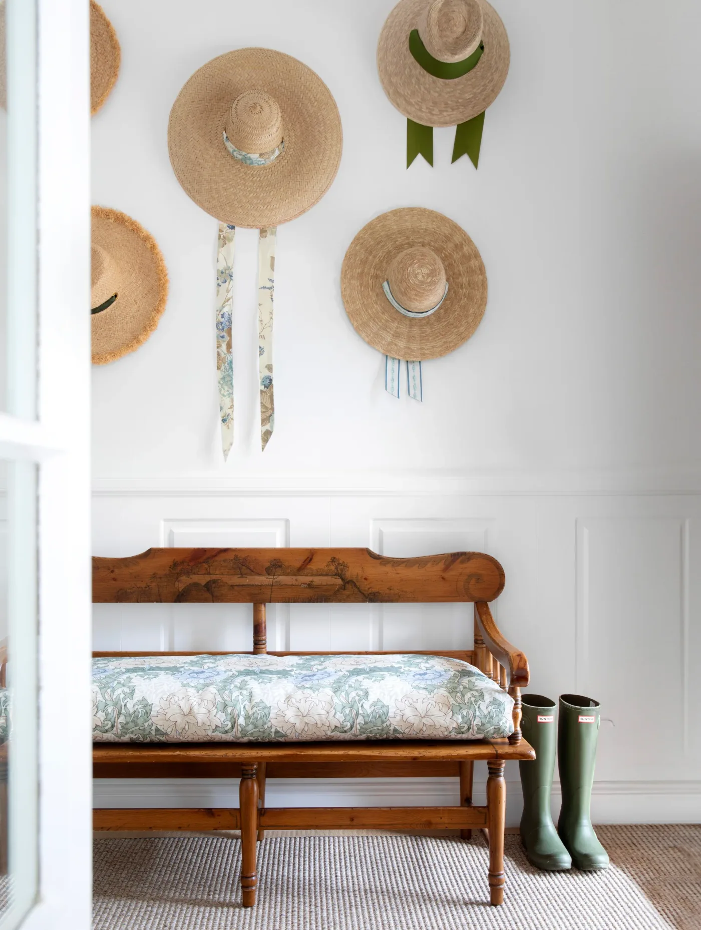 Entry corner with a wooden bench, pale floral cushion, white lower paneling, hanging straw hats and tall boots beside the seat.
