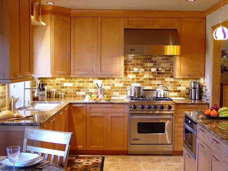 Warm wood framed cabinets surround a broad metal range and hood beside darker small backsplash tiles and a compact adjoining dining surface