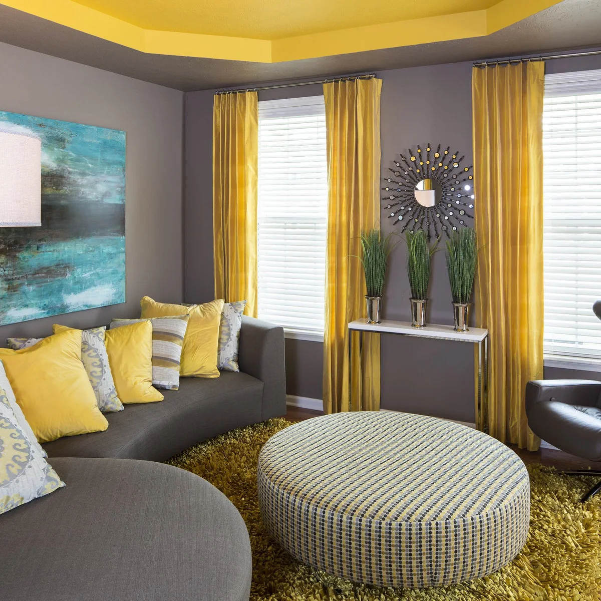 Gray living room with yellow curtains and cushions, a round patterned ottoman and turquoise wall artwork