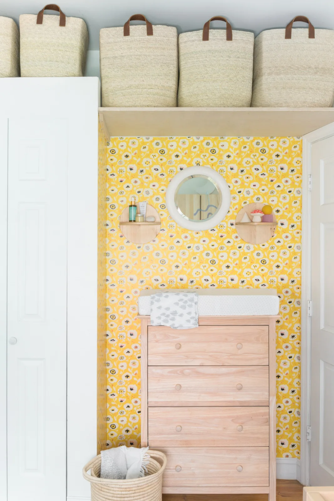 A timber changing chest stands in a yellow floral-wallpapered nook beneath a mirror, small shelves and high storage baskets.