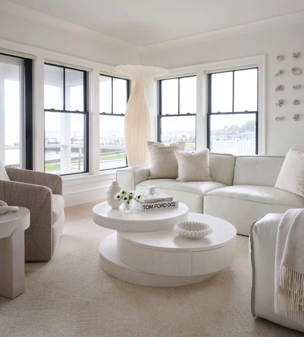 White living room with a curved pale sofa, two rounded nesting tables, soft cushions and dark-framed windows.