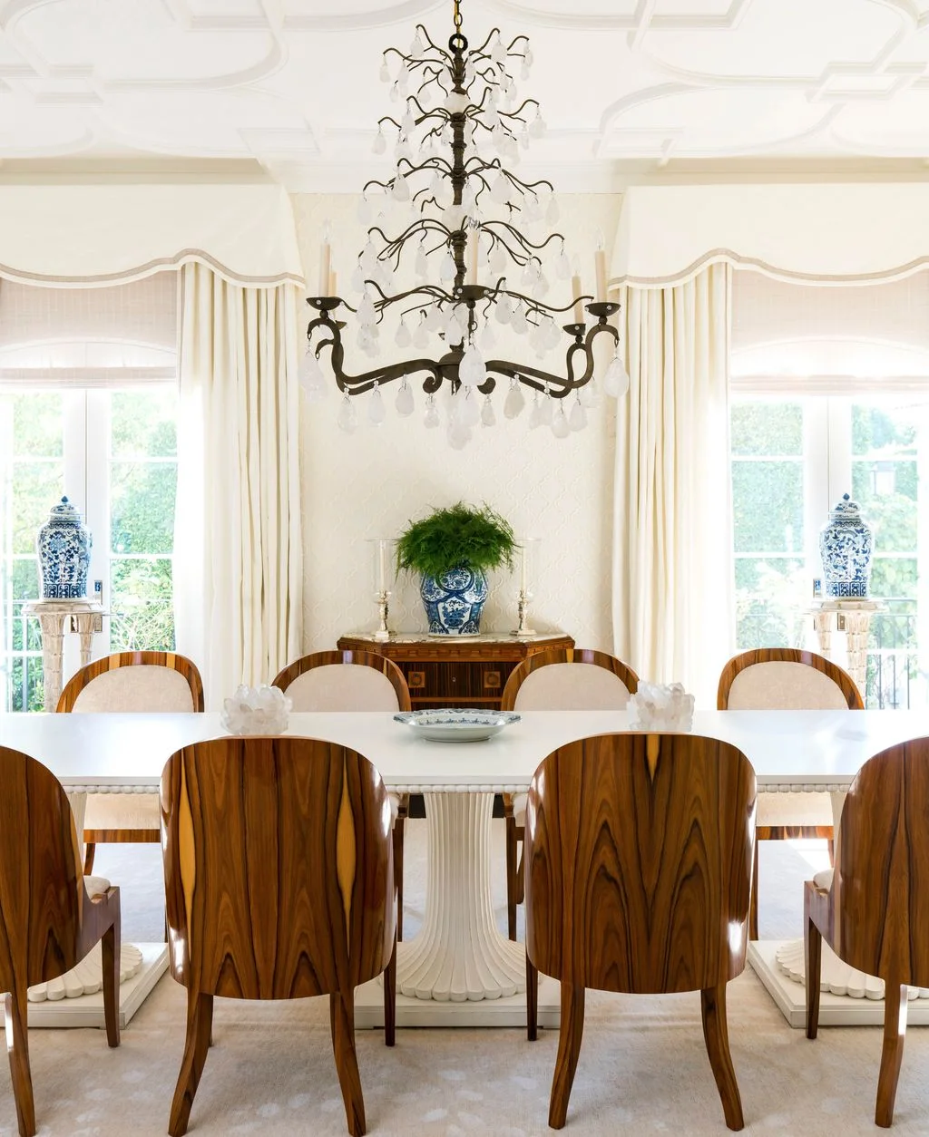 Wooden chairs with strongly patterned backs surround a pale rectangular dining table beneath a tiered dark chandelier beside pale curtains