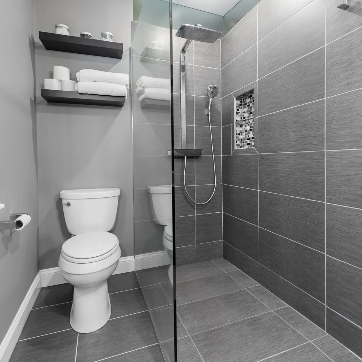 Grey compact bathroom with two dark shelves above the toilet beside a clear shower screen