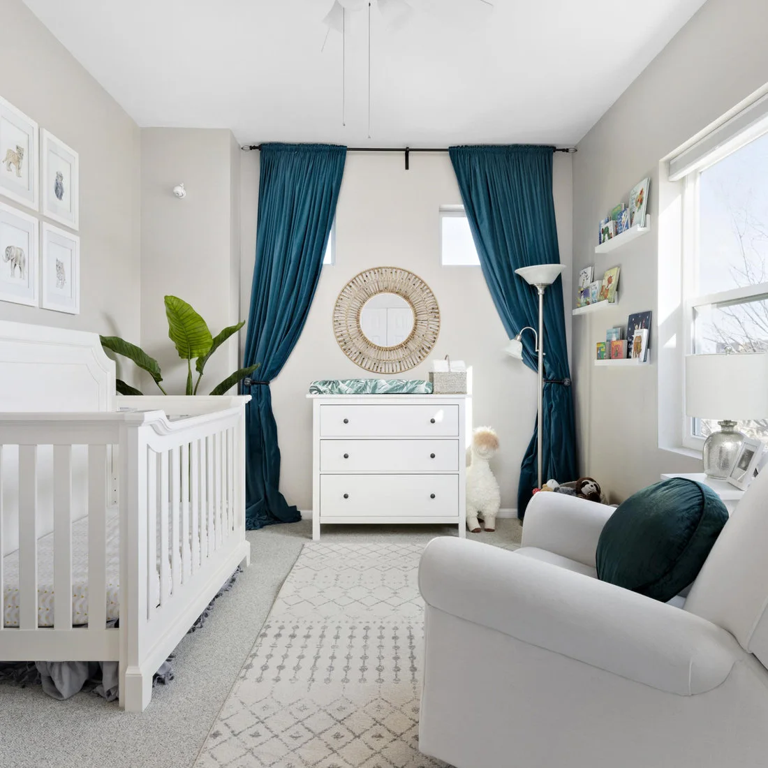 A white crib and changing dresser share a pale room with teal curtains, a round woven mirror and a light chair with a teal cushion.