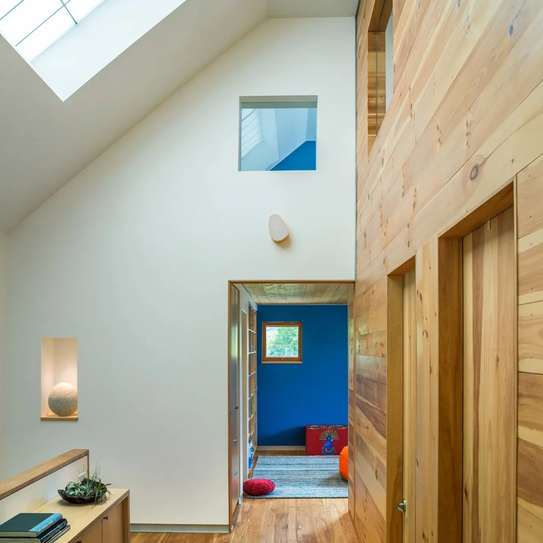 A skylit landing combines a high white wall, pale horizontal wood boards and a doorway looking into a blue room.