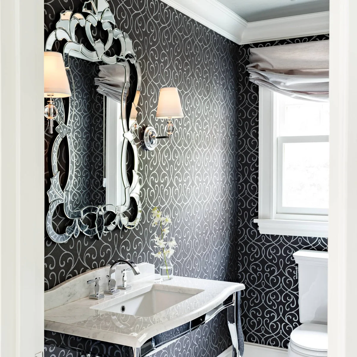 An ornate mirrored frame hangs over a white console basin against dark wallpaper with a pale scrolling pattern.