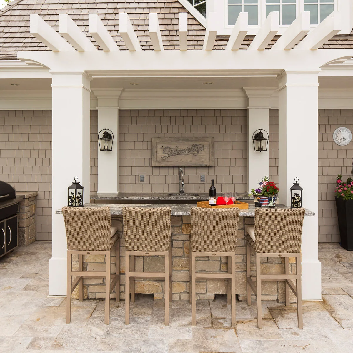 Stone-faced outdoor bar with four woven high-backed chairs between white columns and a sink behind