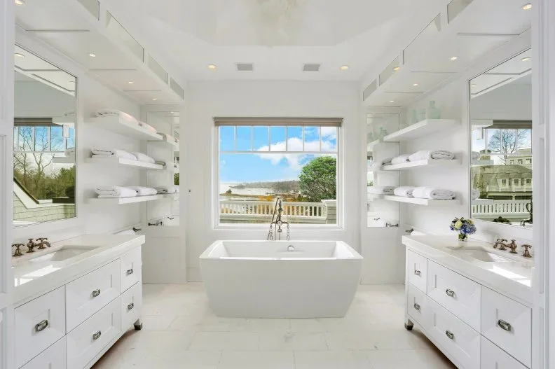 A large white bathroom has a rectangular tub beneath a central window between two vanity runs and separate open shelves holding towels