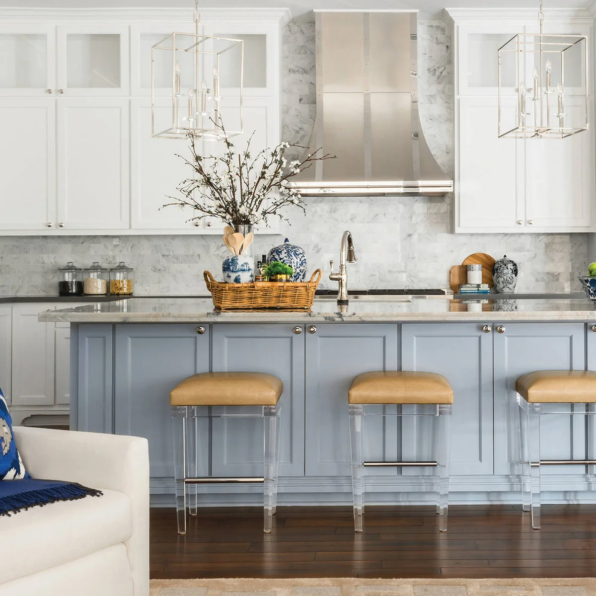 A pale blue island stands below white cabinets and veined pale tile, with ochre padded stools and blue and white accessories.