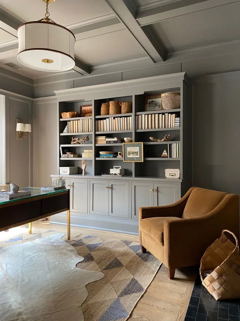 Gray built-in office shelves and a coffered ceiling beside a desk with warm metal legs and a brown armchair.