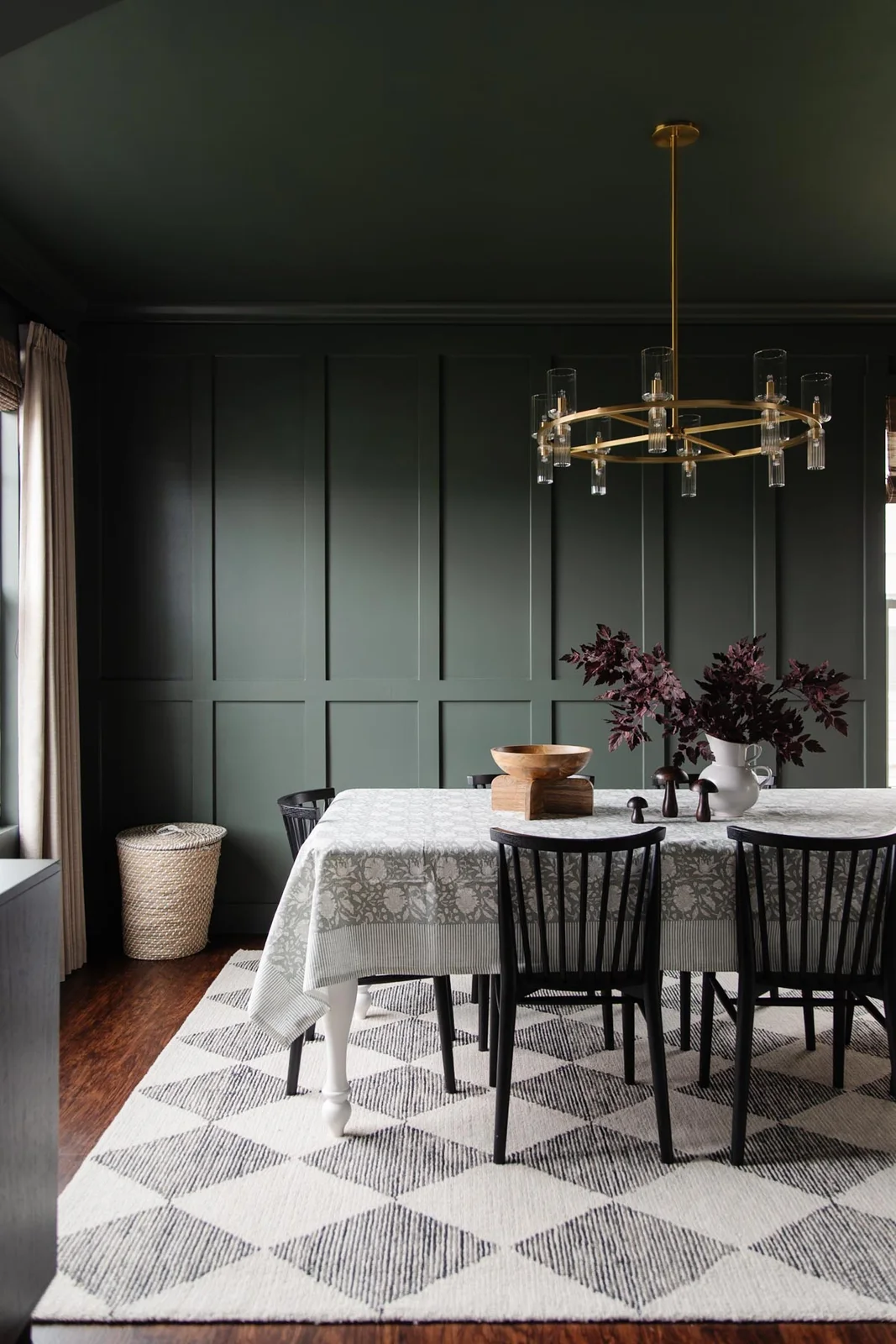 Deep green walls and ceiling surround black spindle chairs, a pale patterned tablecloth and a diamond-patterned rug beneath a circular chandelier