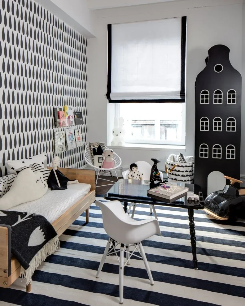 A black-and-white room has teardrop wallpaper, a wood-framed daybed, a low dark table, a white chair, a striped rug and a tall house-shaped chalkboard.