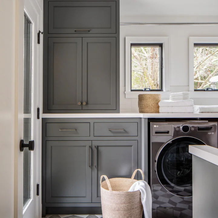 Gray utility cabinets stand beneath corner windows beside woven baskets and patterned floor tiles, with a dark appliance at one side.