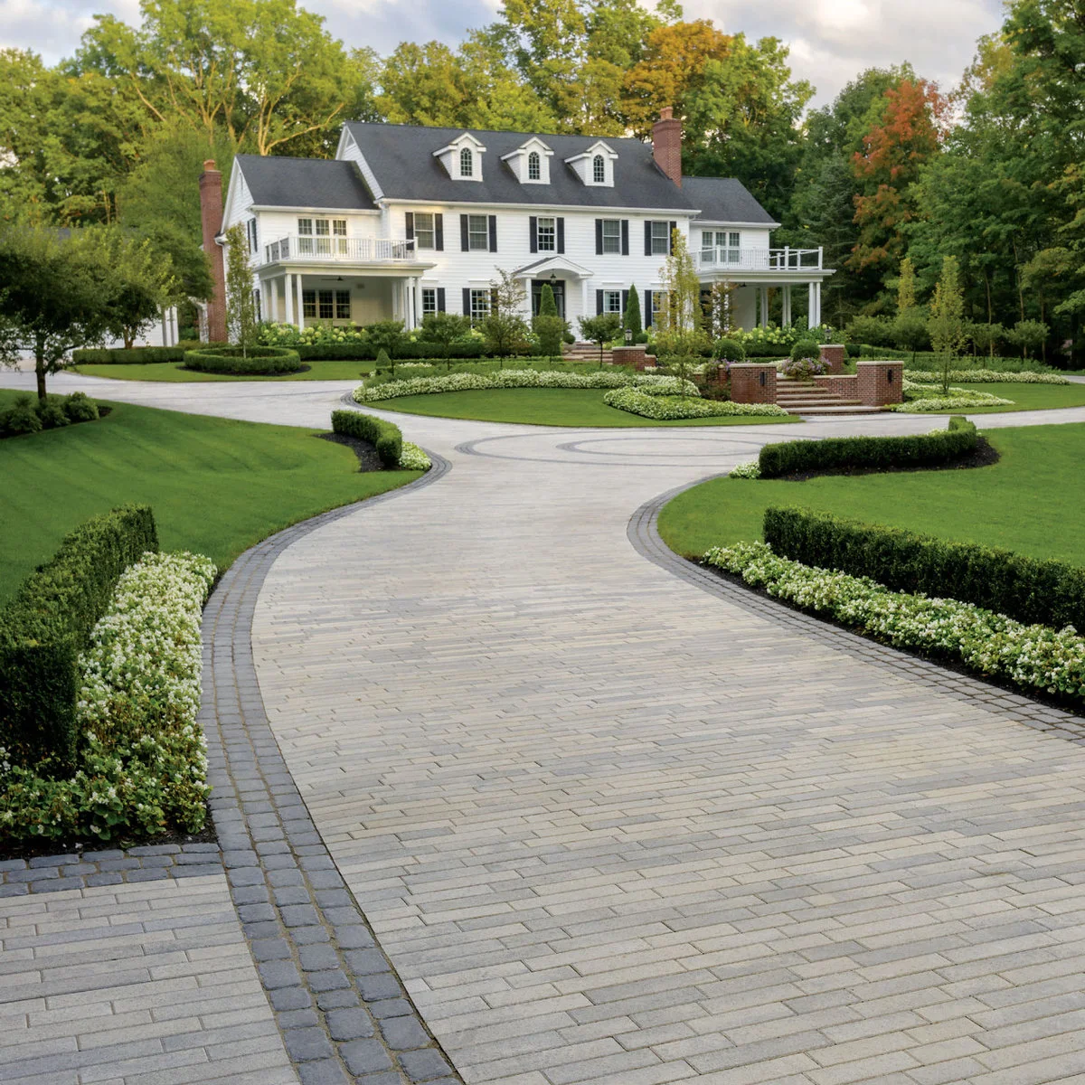 Broad pale paved driveway curving around a planted central island in front of a white house.