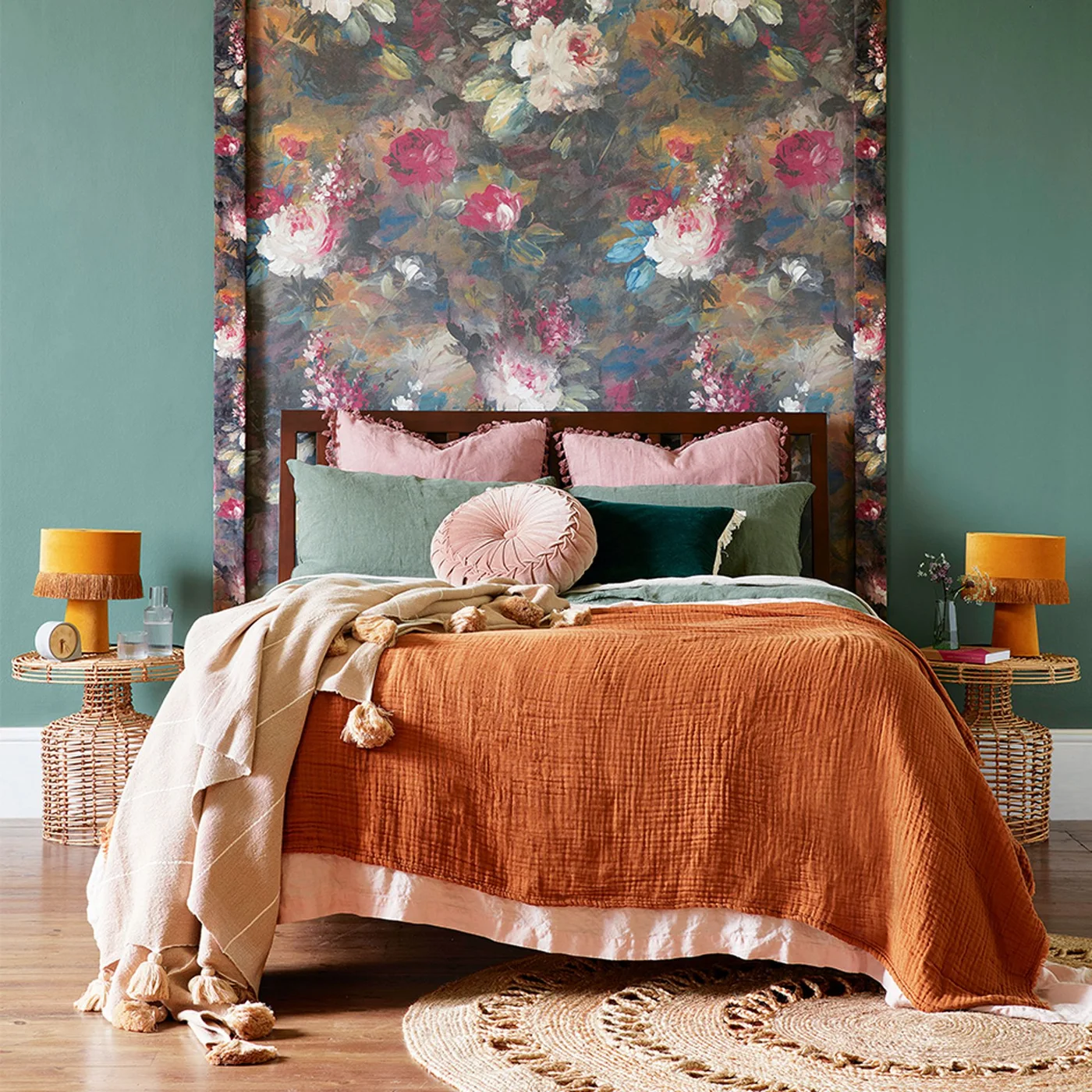 Green walls frame a floral panel behind a wooden bed with pink cushions, an orange throw and woven bedside tables