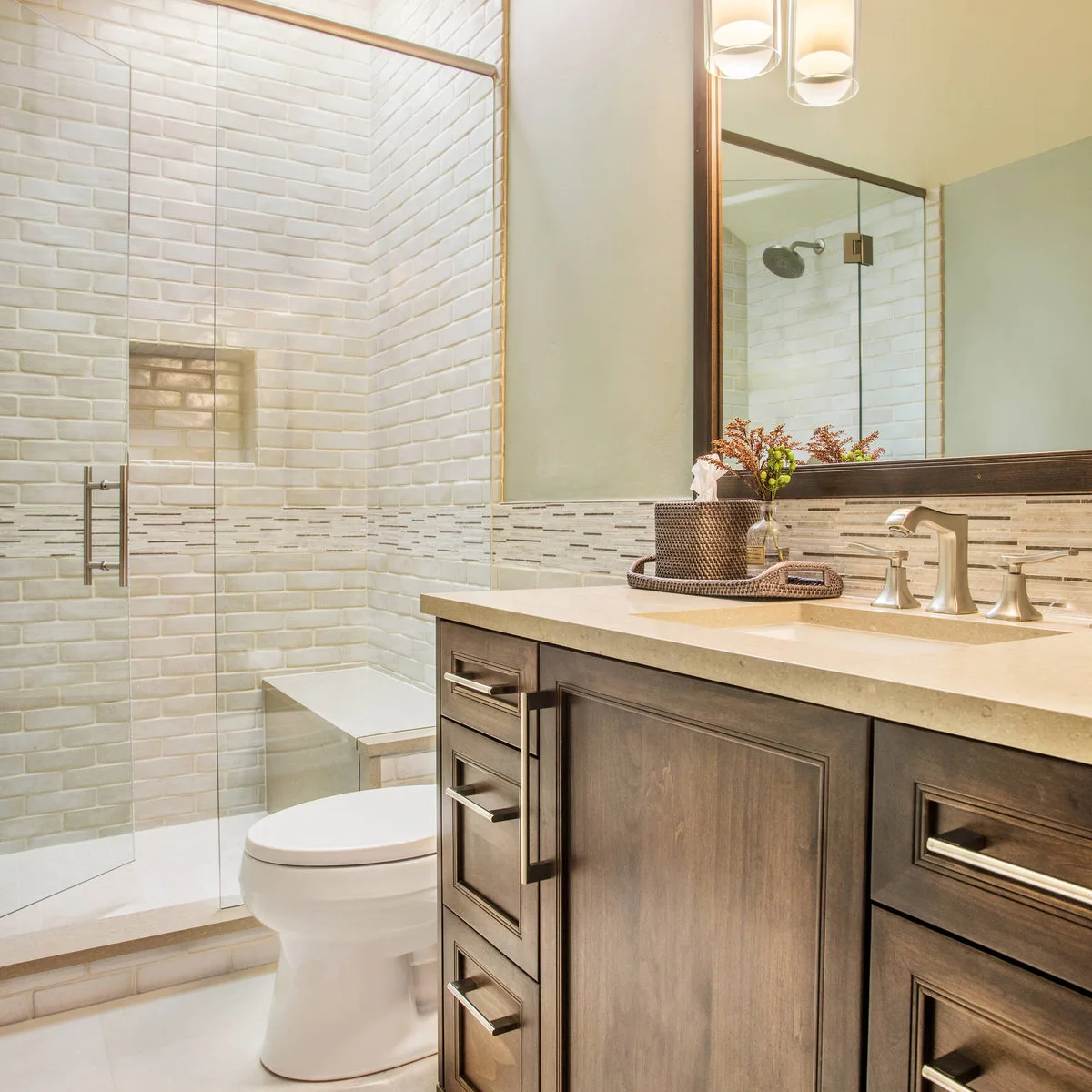A glass shower with softly varied white brick-shaped tiles, a recessed niche and bench beside a dark wood vanity.