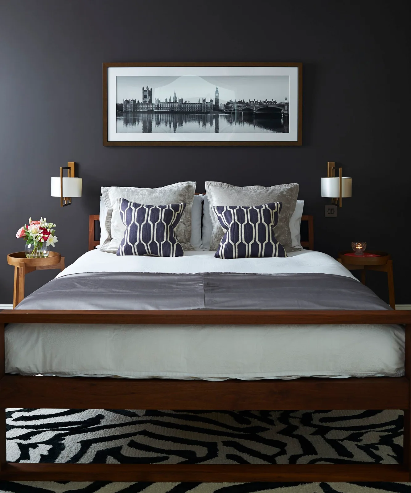 White and gray bedding sits beneath a framed horizontal photograph on a charcoal wall with two wall lamps and a patterned rug