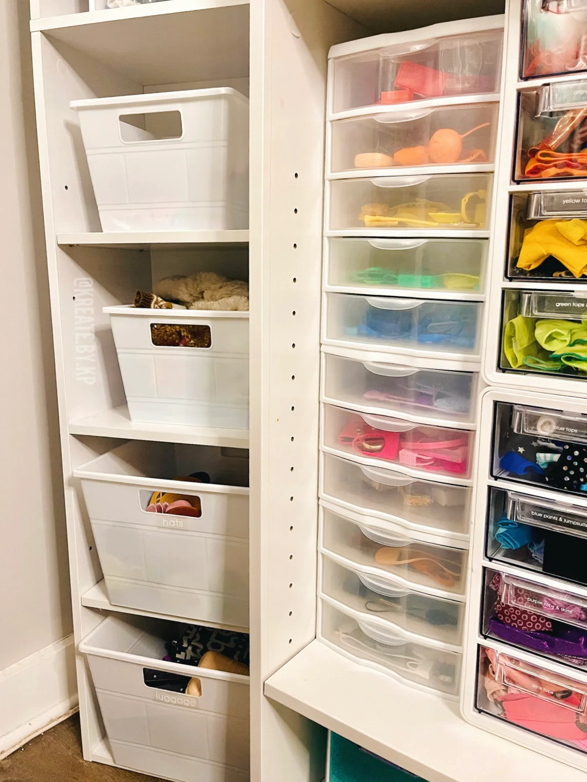 Closet shelving with white bins for hats and luggage beside clear shallow drawers holding color-sorted doll clothes and accessories.