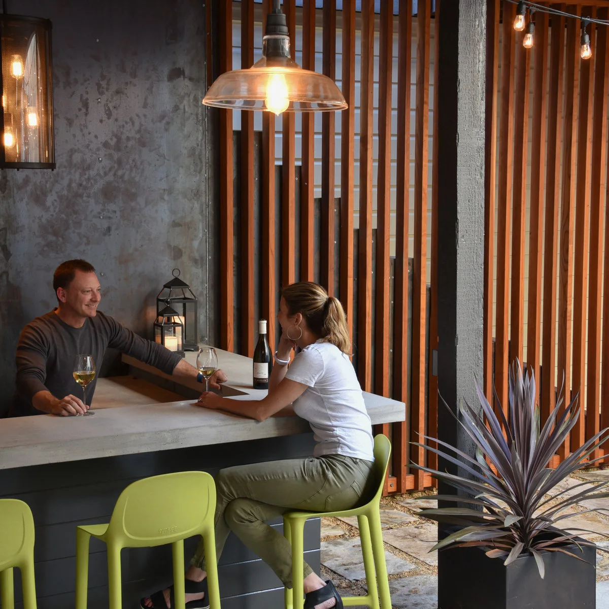 Small dark outdoor bar with a pale counter, lime green stools and vertical timber screening beside a potted plant
