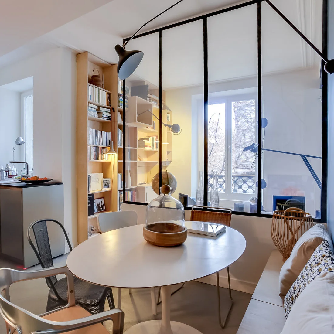 A round white dining table and mixed chairs sit beside a cushioned bench and a black-framed glazed partition.