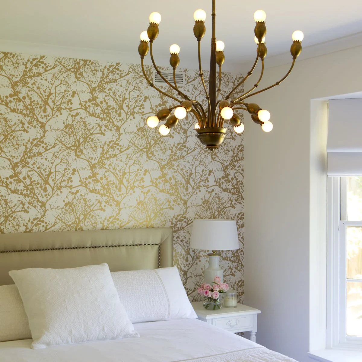 Gold-colored branch wallpaper behind a pale upholstered headboard with white bedding and a branching chandelier.