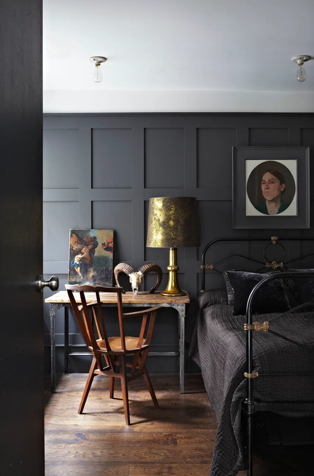 A dark paneled bedroom has a black open bed frame beside a wood desk, chair and gold-colored table lamp beneath a white ceiling
