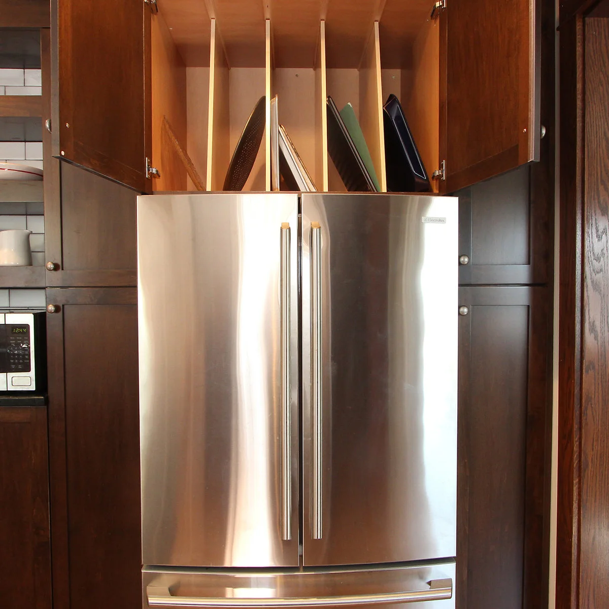 Vertical dividers separate baking trays and boards inside a dark cabinet above a French-door refrigerator.