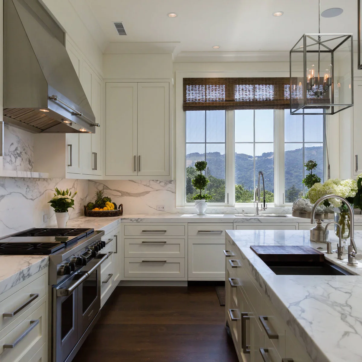 A woven blind raised above a wide kitchen sink window with white cabinets and pale veined counters.