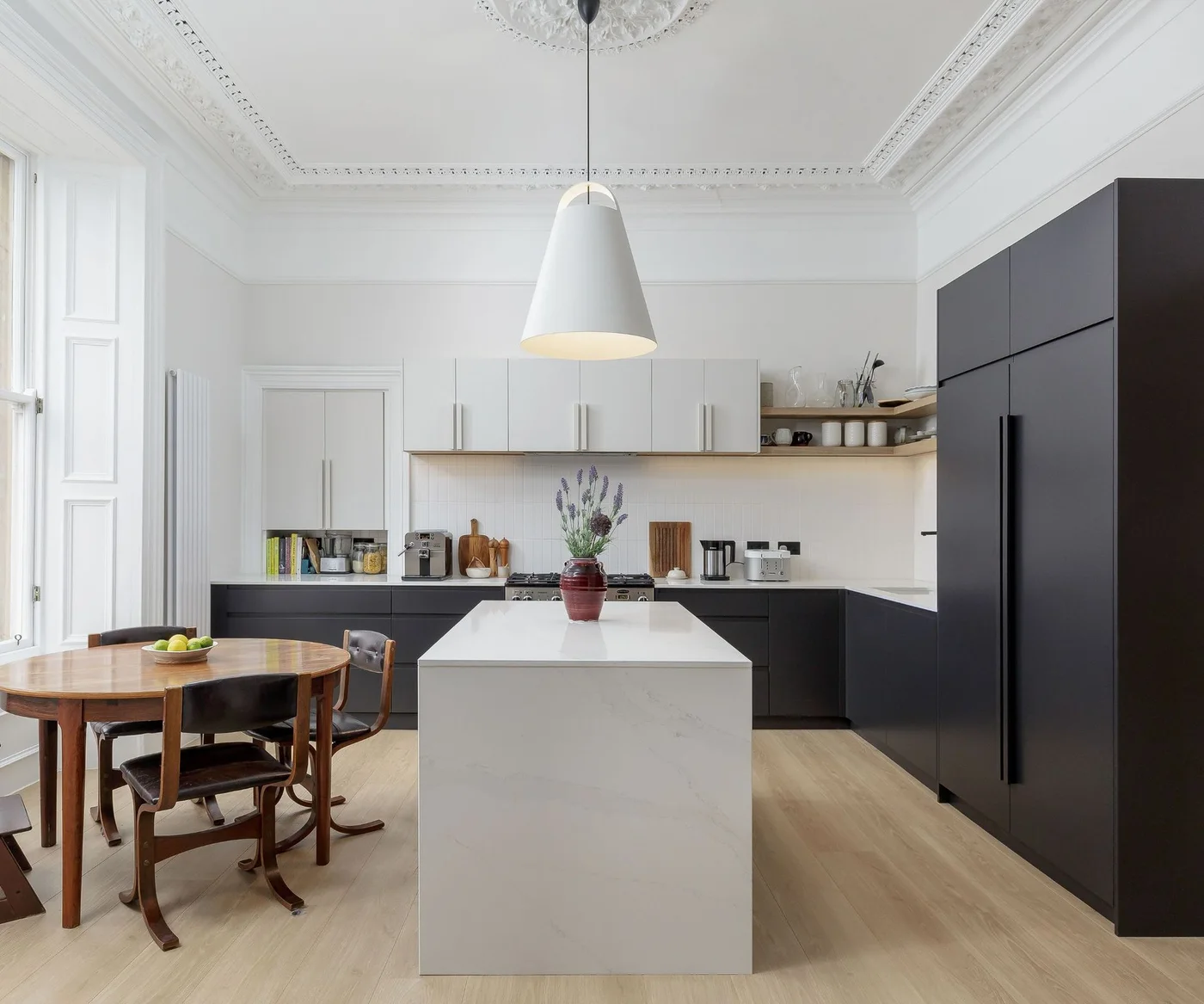 Dark flat-front lower and tall cabinets face a white waterfall island, with white uppers, open corner shelves and a round dining table.