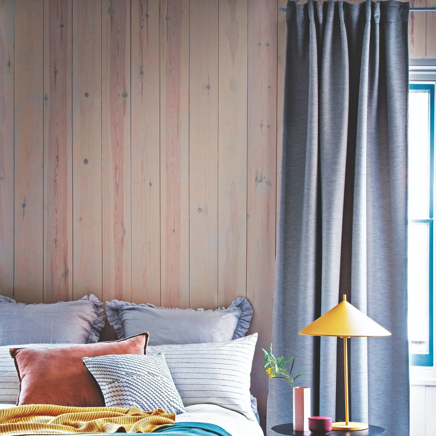 Pale wood wall paneling and long gray curtains frame a bed with gray pillows, a mustard throw, a coral cushion and a yellow bedside lamp.