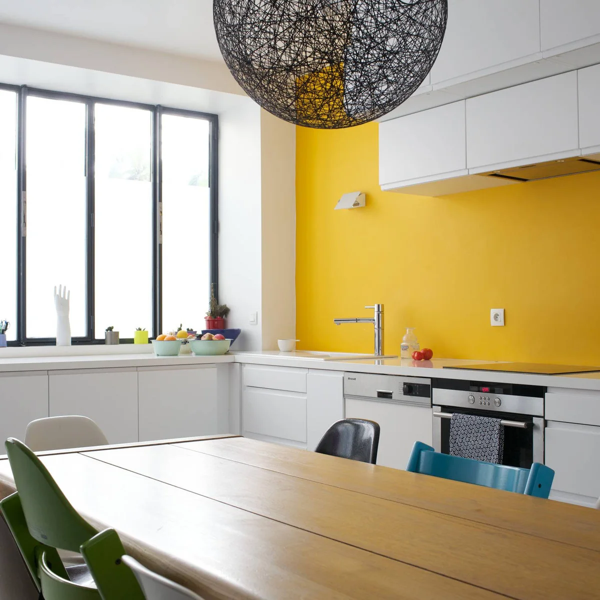 White kitchen cabinets beneath a yellow wall with black window frames and colorful dining chairs