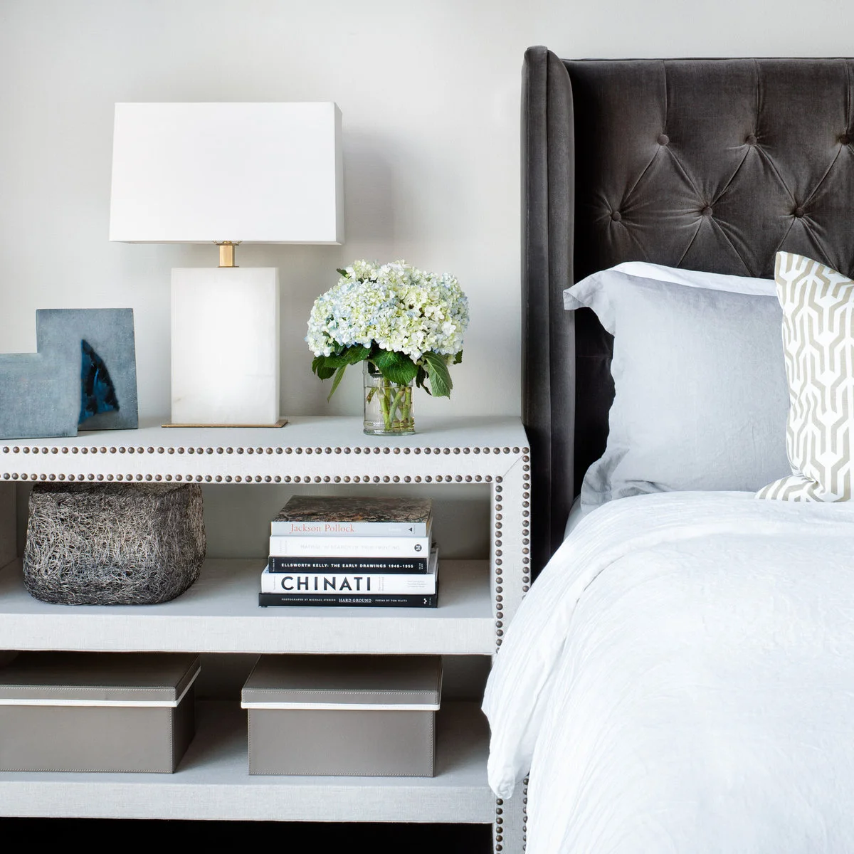 Pale open-shelf nightstand with books, two lidded boxes and nailhead edging