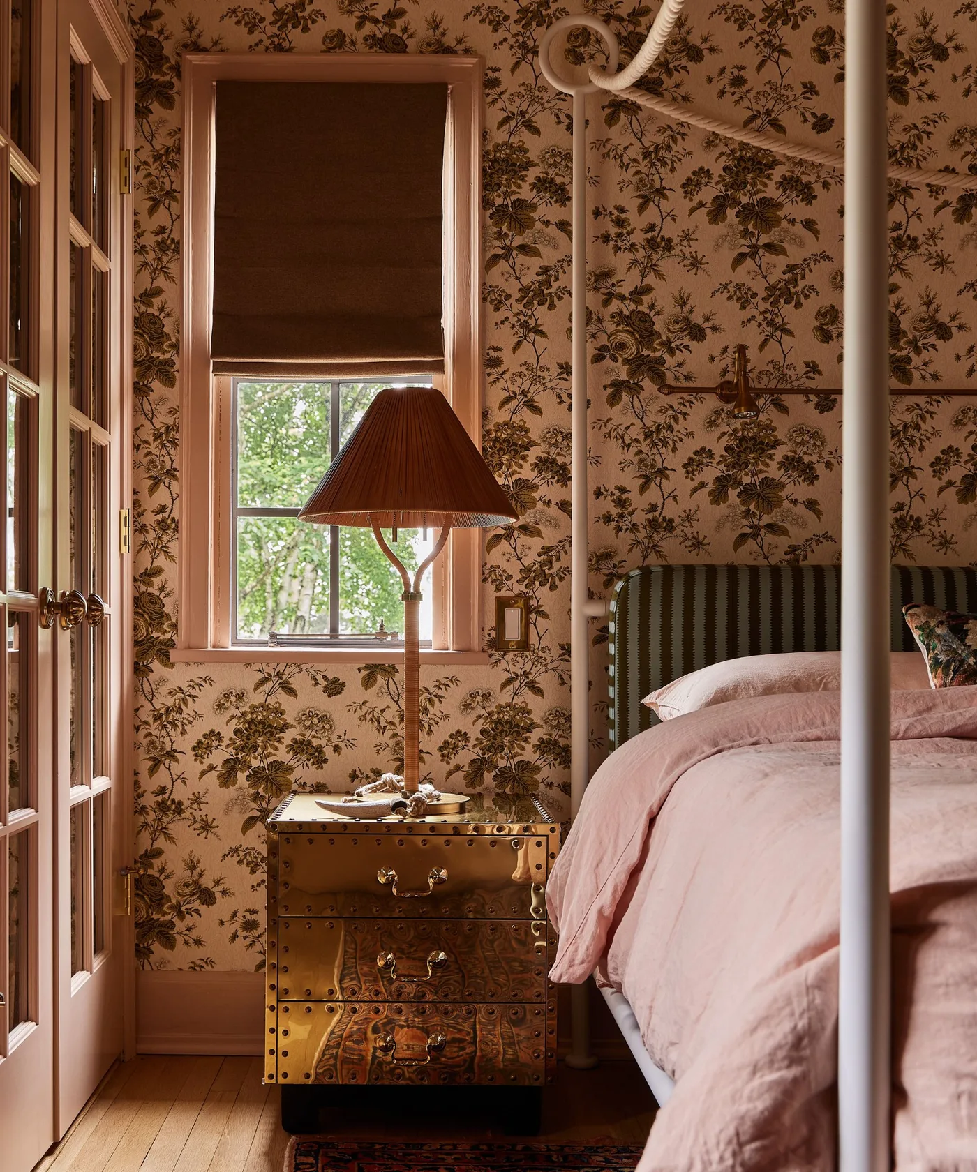 Green-and-brown floral wallpaper behind a gold-toned bedside chest, woven lamp and pale canopy post.