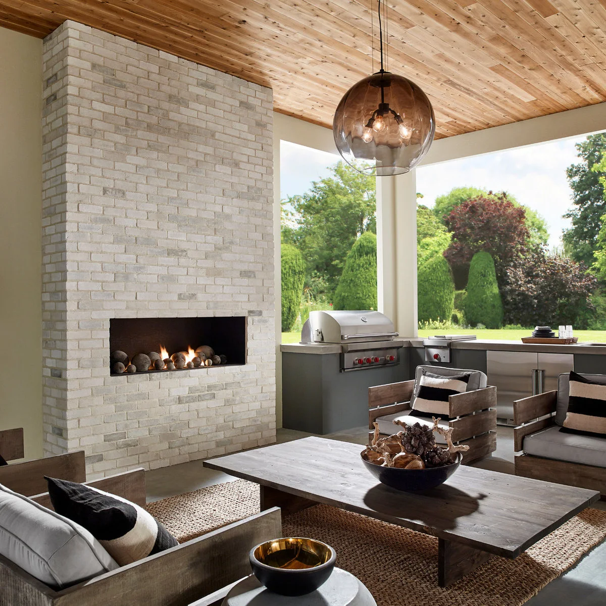 Outdoor sitting area with pale brick fireplace wall, wood-framed chairs and a grill counter beyond