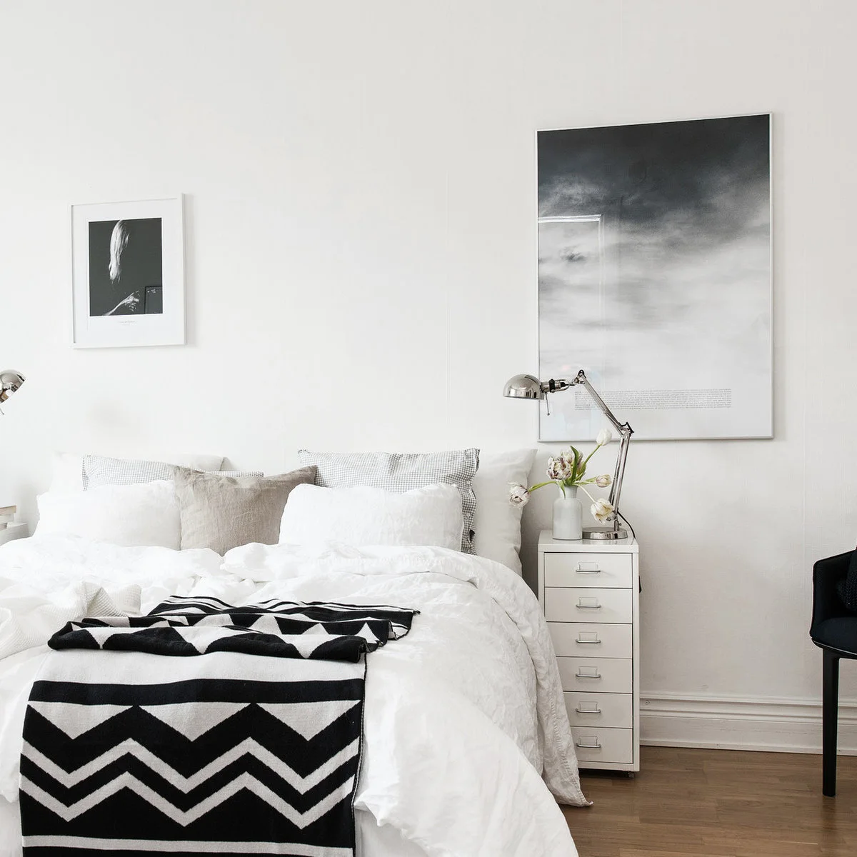 White bedroom with a narrow multi-drawer bedside cabinet, metal task lamp and black patterned throw