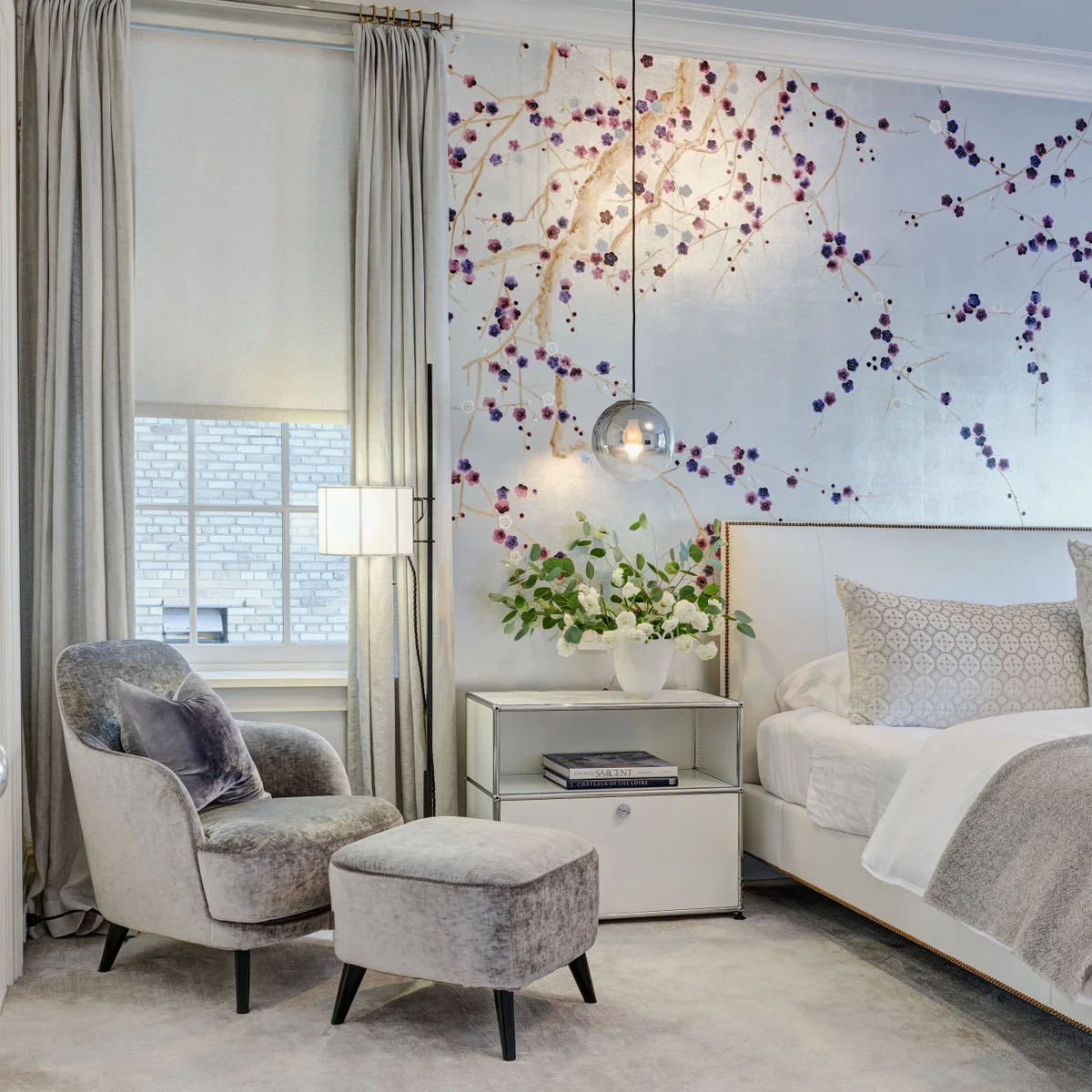 Pale branch-and-blossom wallpaper behind a white bed beside gray curtains, an upholstered chair and a silver pendant.