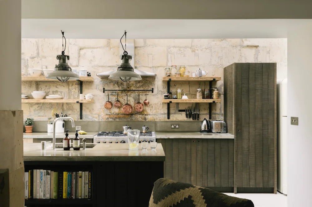 Rustic kitchen with deep olive-gray paneled cabinets, tall cupboard, dark island with book shelf, pale textured wall, wooden shelves, and hanging pots.
