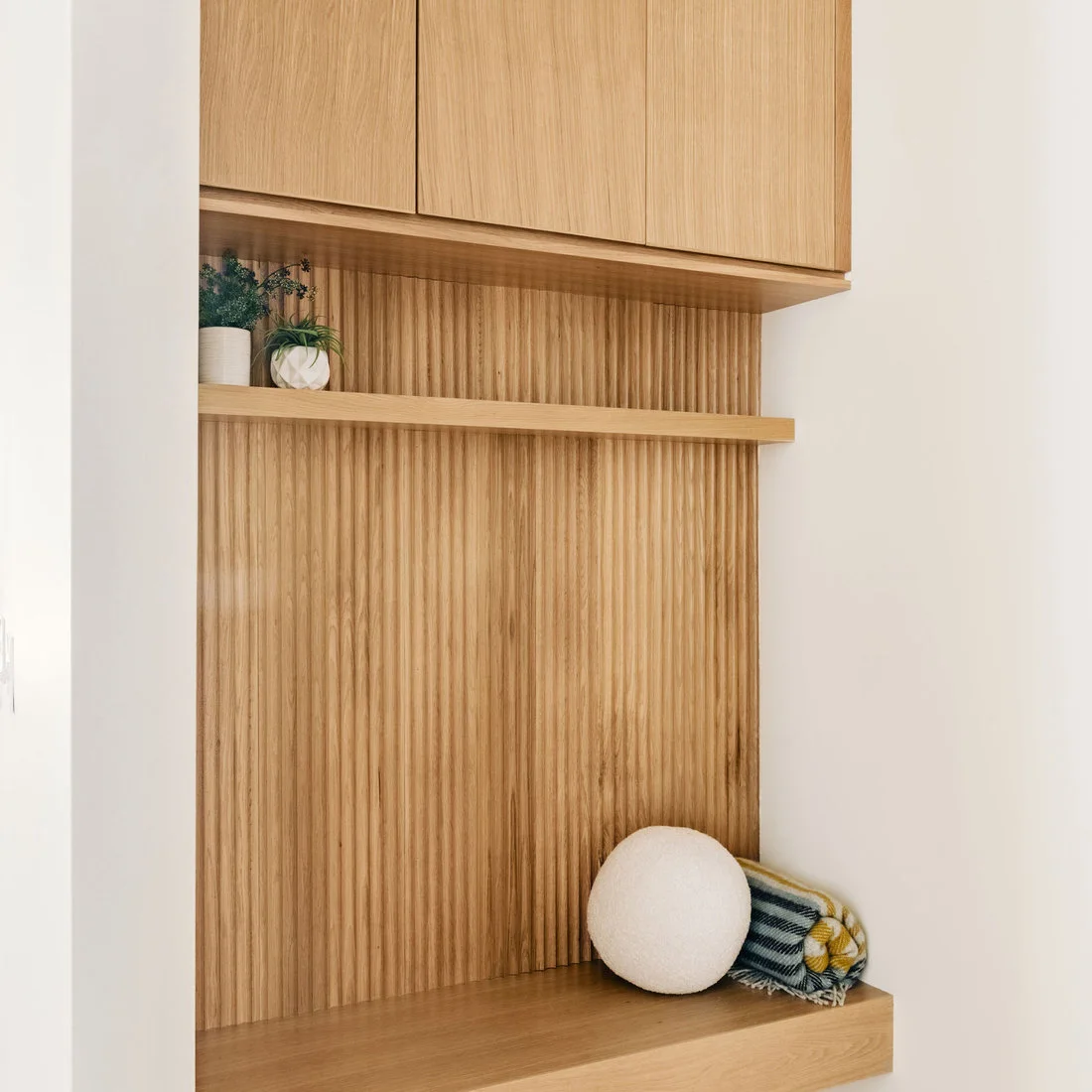 A compact wood bench sits below a wood shelf and overhead cupboards against a vertical wood back panel with a pale cushion.