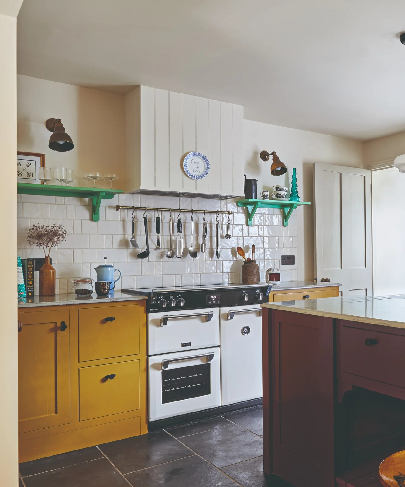 Mustard base cabinets and a darker island face a white cooker beneath green shelves, hanging utensils and a plain pale hood.
